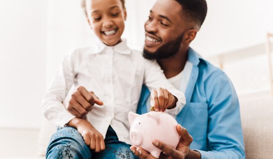 Girl and father putting coin into piggy bank, sitting on sofa at home